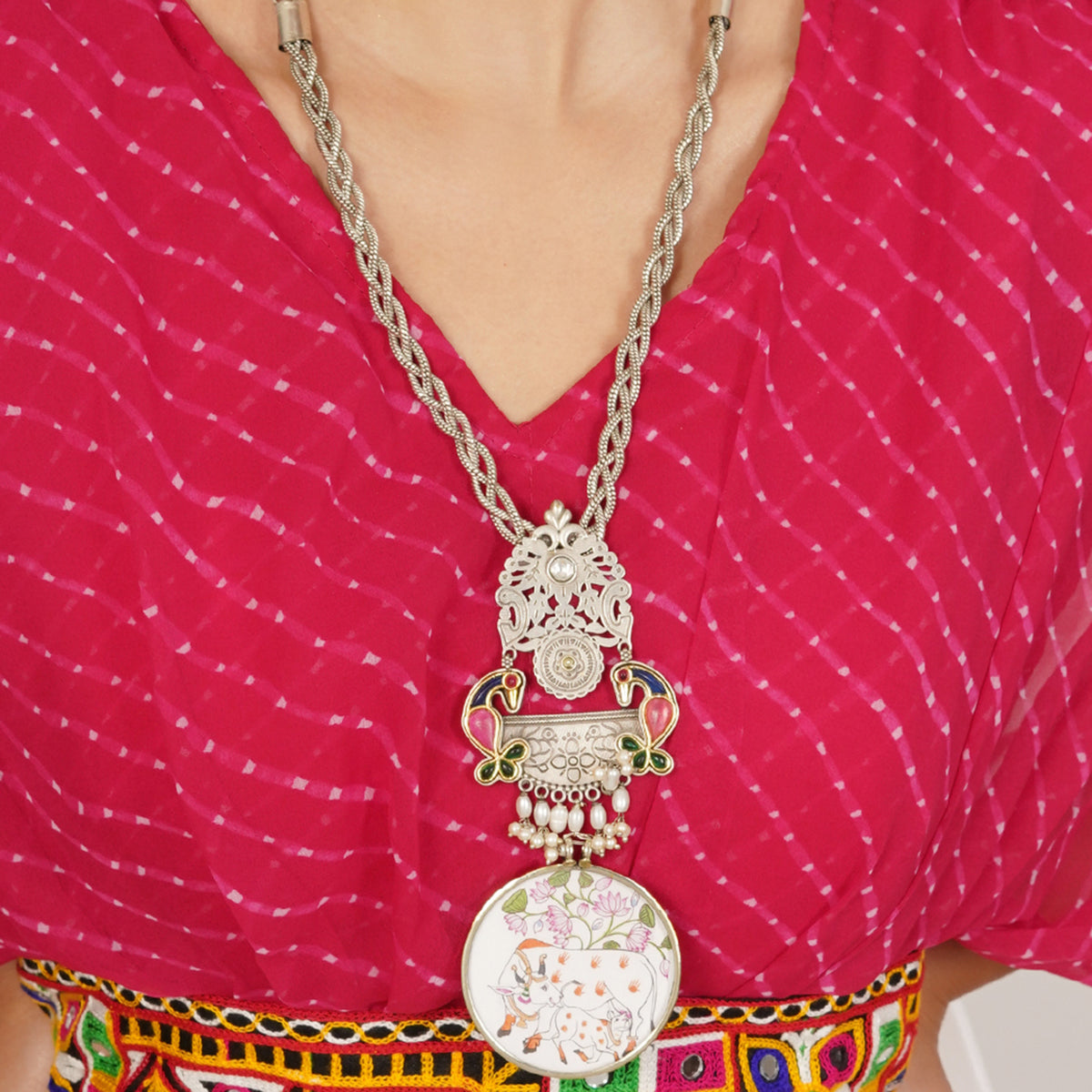 A close-up of a person wearing a large ornate silver-toned pendant necklace with floral and paisley details over a bright pink patterned top.