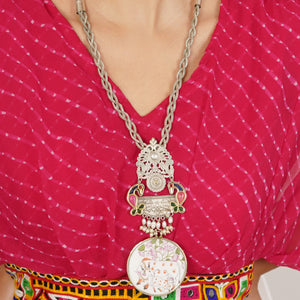A close-up of a person wearing a large ornate silver-toned pendant necklace with floral and paisley details over a bright pink patterned top.
