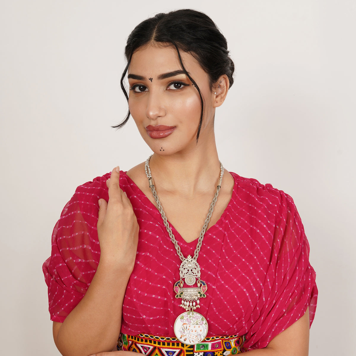 A woman wearing a bright pink tie-dye blouse and a colorful patterned belt, accessorized with a large silver pendant necklace, photographed against a plain light background.