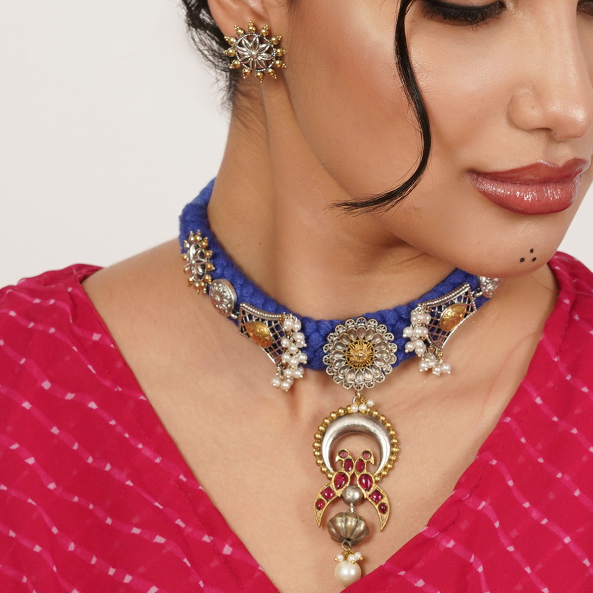A close-up of a woman wearing an ornate royal blue choker necklace with gold and silver-toned embellishments, white pearls, and a large crescent-shaped pendant with red stones and a pearl drop.