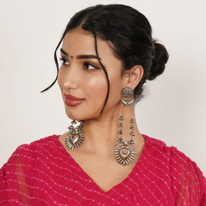 A woman wearing large, ornate silver-toned dangle earrings with sunburst and crescent moon motifs.
