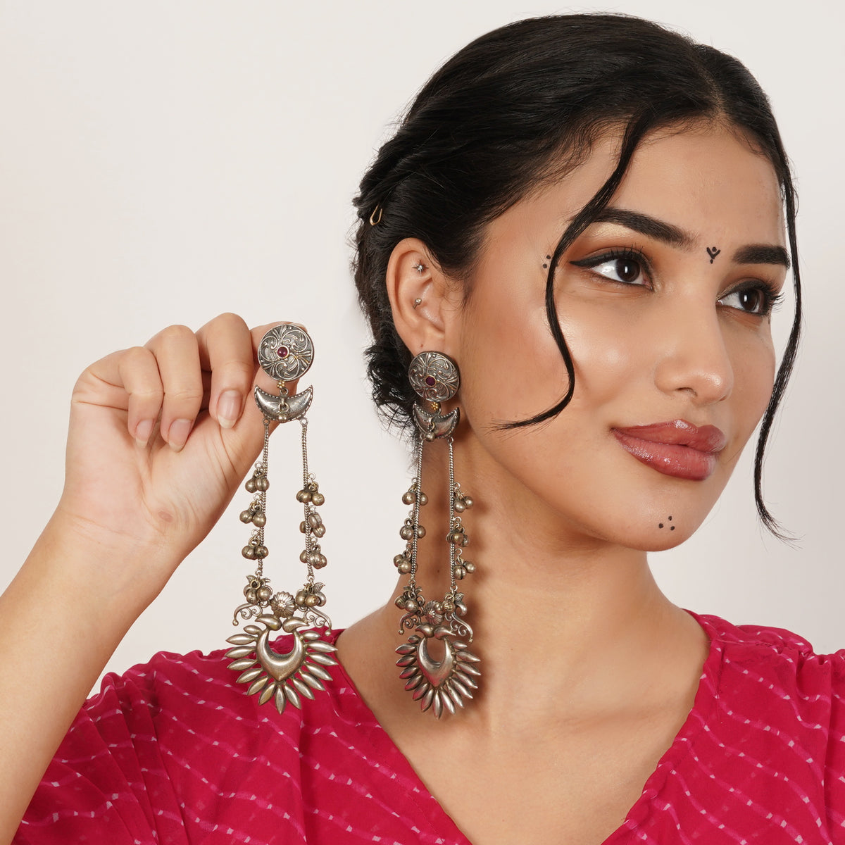 A woman wearing traditional Indian jewelry, holding up a pair of silver-toned, multi-strand dangle earrings with small pink gemstone accents.