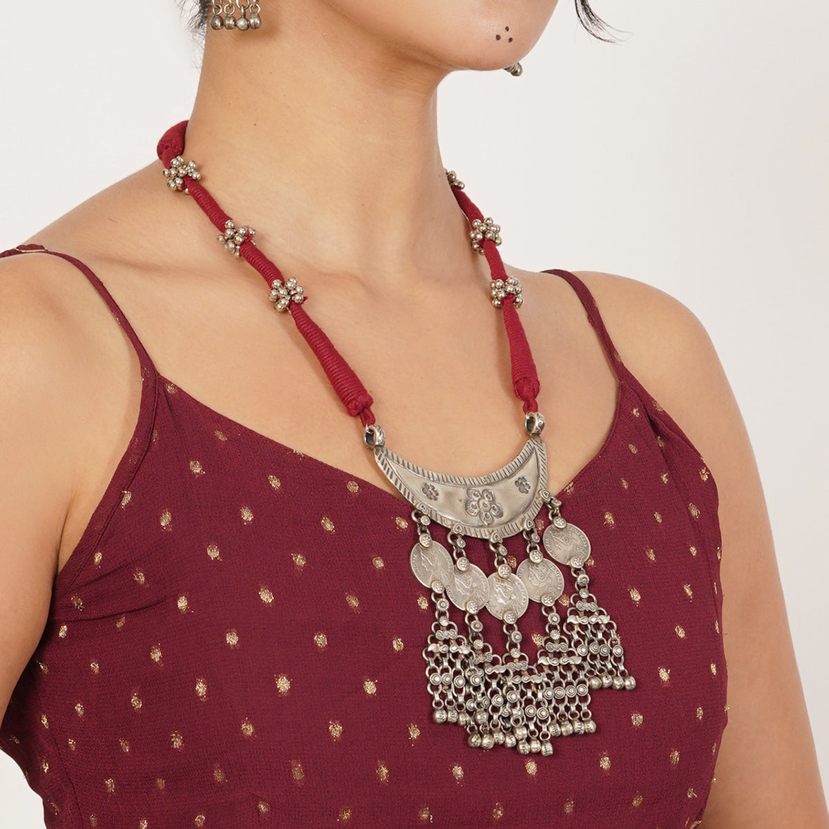 A close-up of a model wearing a traditional silver coin pendant necklace with a red cord and dangling silver bells, paired with a maroon polka-dot top.