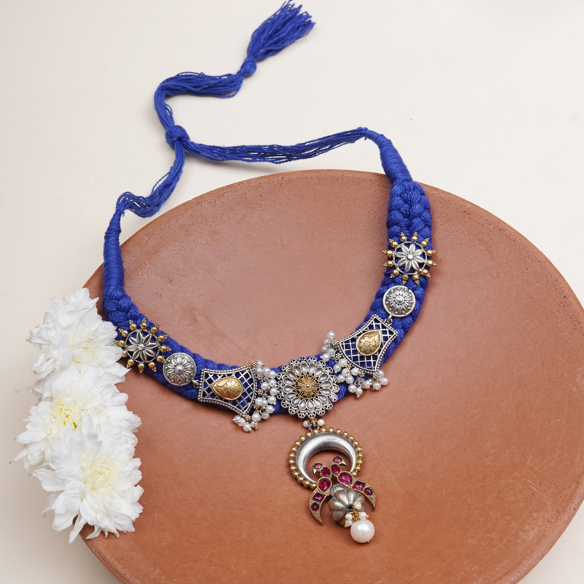 A royal blue braided necklace with ornate silver-toned and gold-toned metal embellishments, white pearl-like beads, and a dangling decorative pendant.