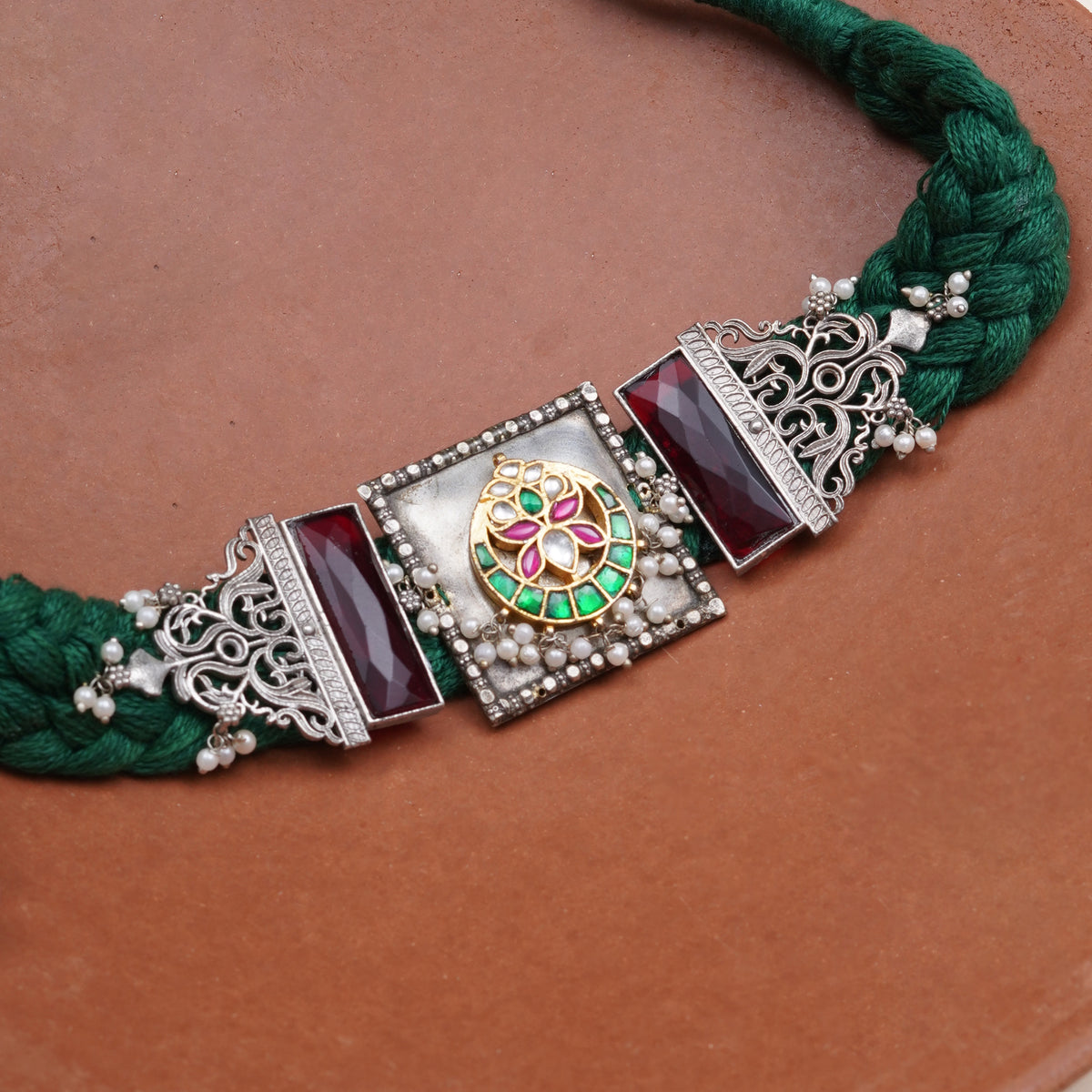 A close-up of a traditional Indian jewelry necklace featuring a dark green braided cord, ornate silver filigree, deep red stones, and a central gold-toned medallion with green and pink enamel details