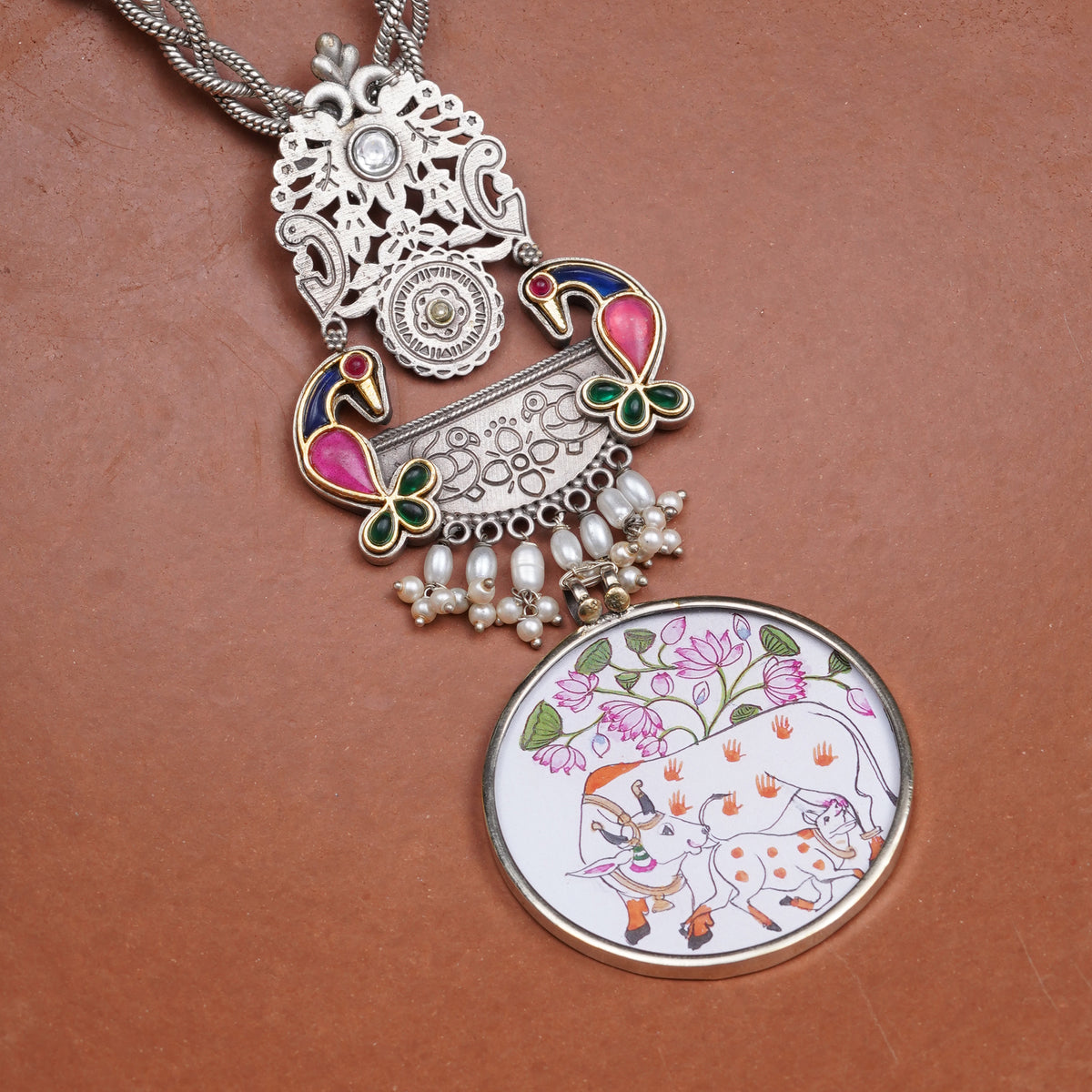 An ornate silver-toned pendant necklace with intricate filigree, colorful enamel details, and a round illustrated pendant featuring a white cow with pink flowers