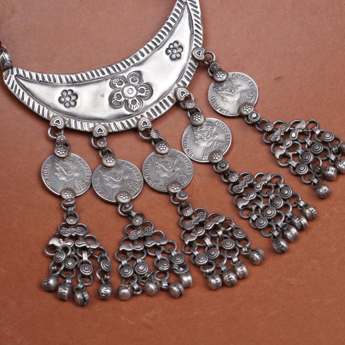 An ornate silver-toned traditional necklace featuring a crescent-shaped collar with engraved floral motifs and five dangling coin-and-fleur-de-lis tassel charms