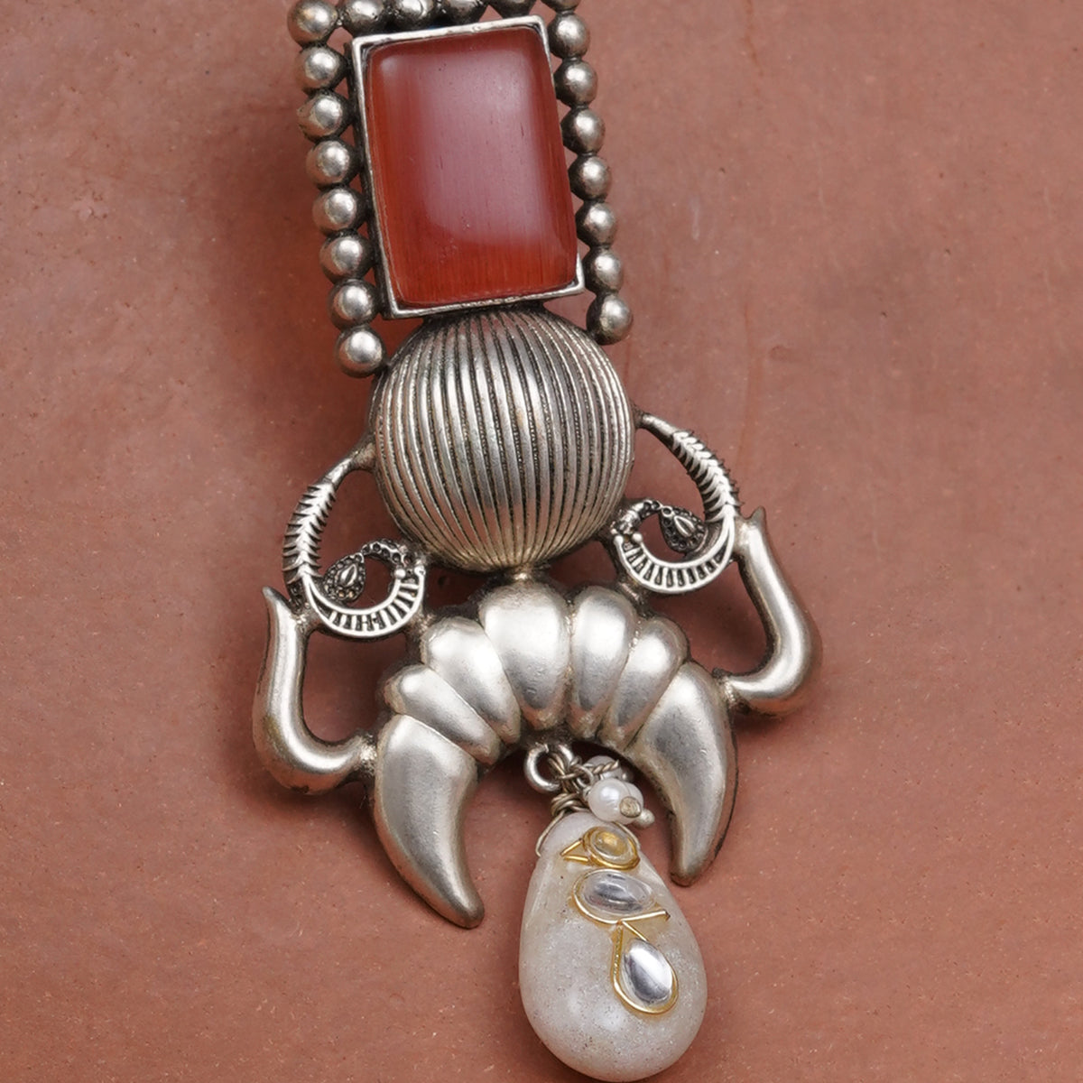 A close-up of a silver-toned jewelry earring featuring a rectangular red stone at the top, a ribbed round silver bead, curved silver details, and a dangling teardrop-shaped pale stone with small gold accents