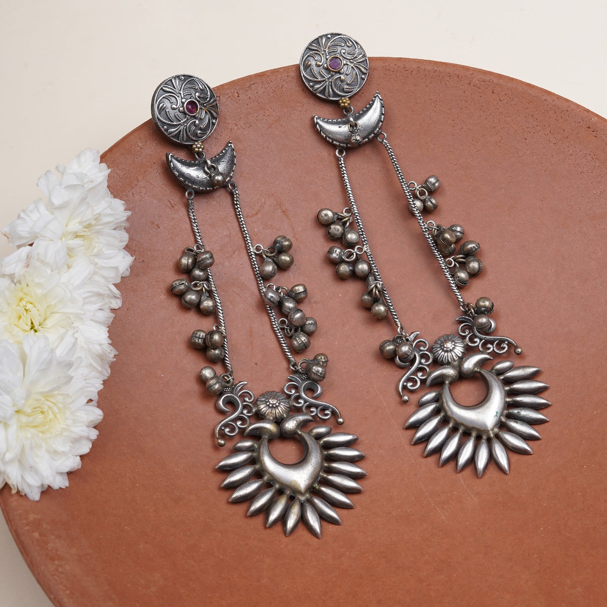 A pair of ornate silver-toned dangle earrings. The earrings feature circular studs with small red gemstone accents, crescent moon charms, beaded chains, and sunburst heart-shaped pendants.