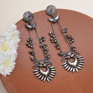 A pair of ornate silver-toned dangle earrings. The earrings feature circular studs with small red gemstone accents, crescent moon charms, beaded chains, and sunburst heart-shaped pendants.