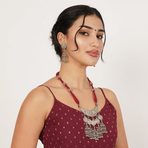 A woman wearing a deep red beaded necklace with a silver-toned ornate pendant and matching dangling earrings
